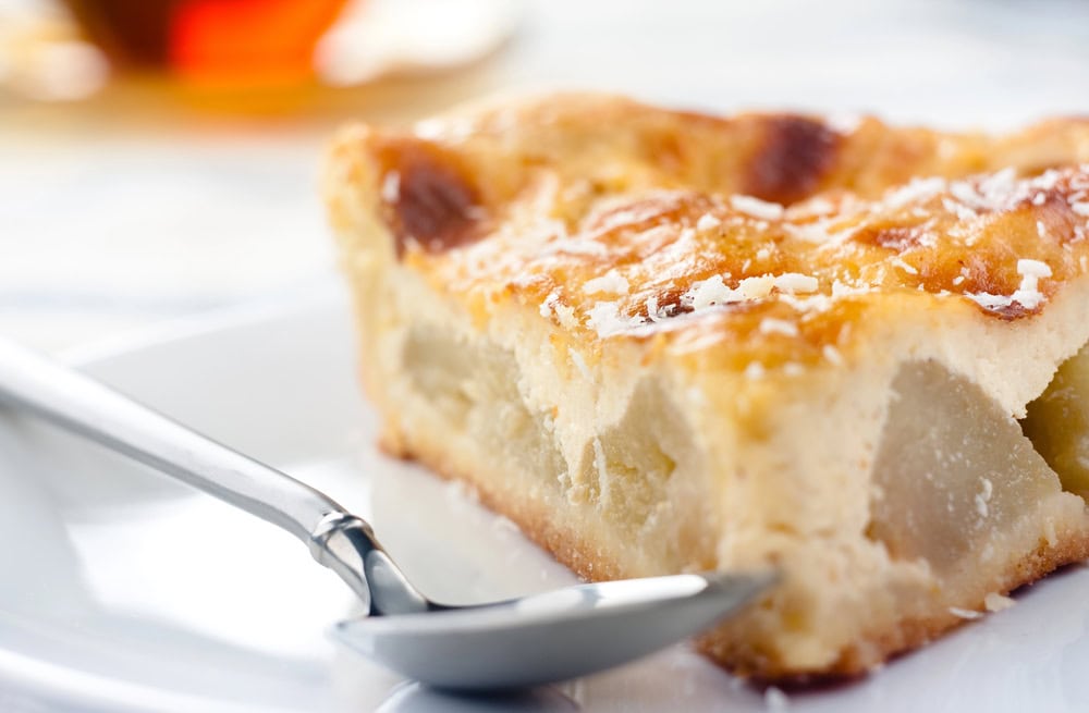 A close-up of a slice of pear tart on a white plate with a spoon beside it.