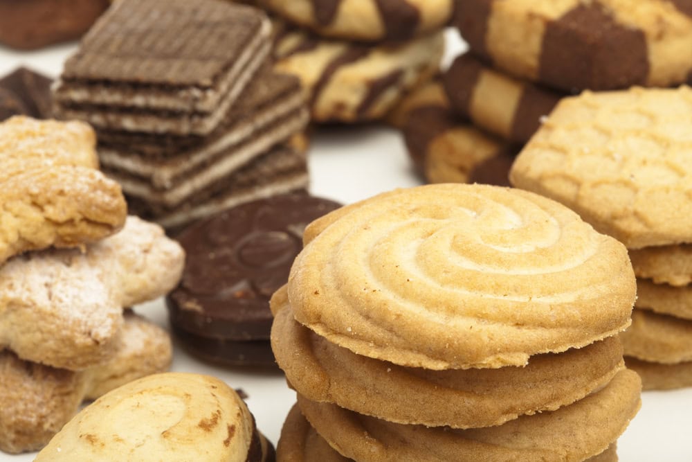 Assorted cookies, including round butter cookies, wafer cookies, and chocolate cookies, arranged in stacks and piles on a white surface.