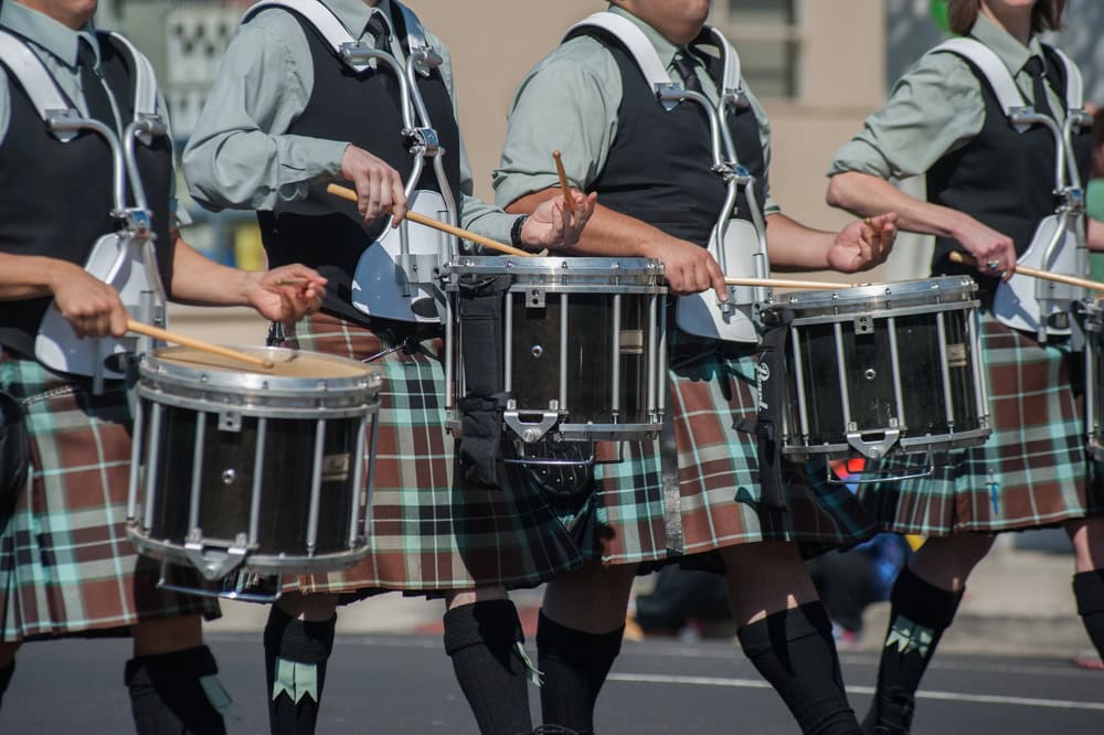 Four people in plaid kilts and uniform shirts play marching snare drums in a parade. Their faces are not visible.