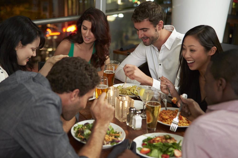 Five people sit around a restaurant table, eating various dishes and drinks, smiling and talking together in a lively atmosphere.
