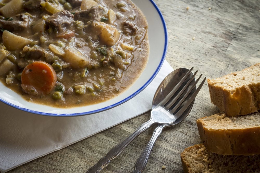 A bowl of hearty stew with vegetables and meat on a plate, accompanied by slices of bread, a fork, and a spoon on a wooden table.