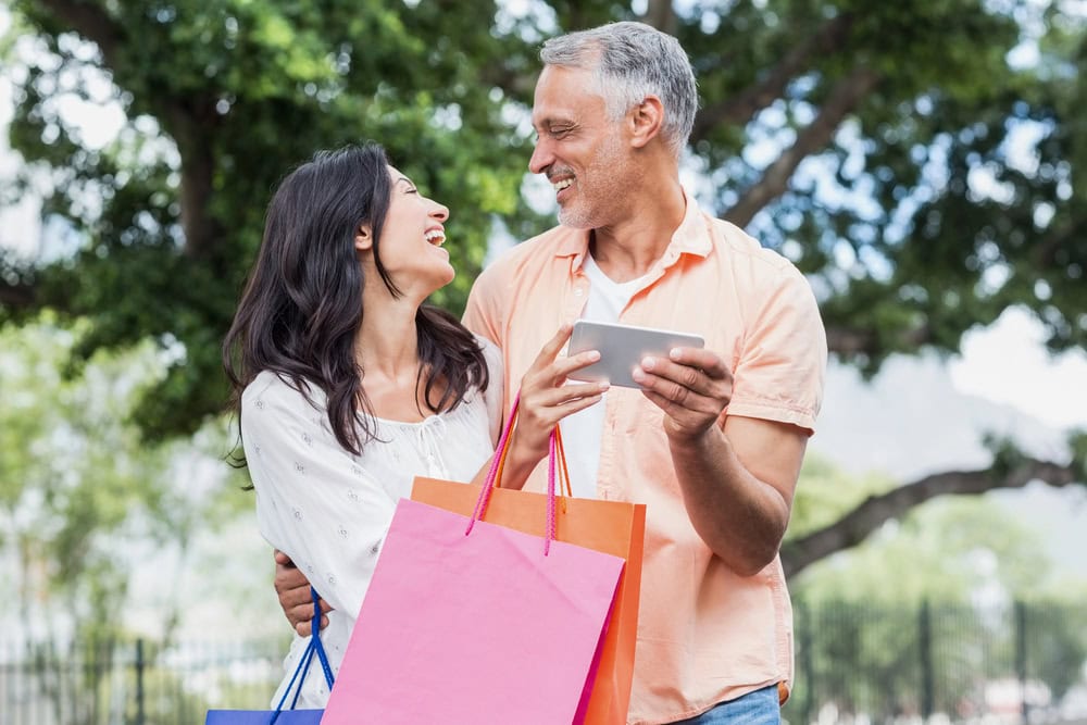 A man and woman outdoors holding shopping bags and a tablet, smiling at each other, with trees in the background.