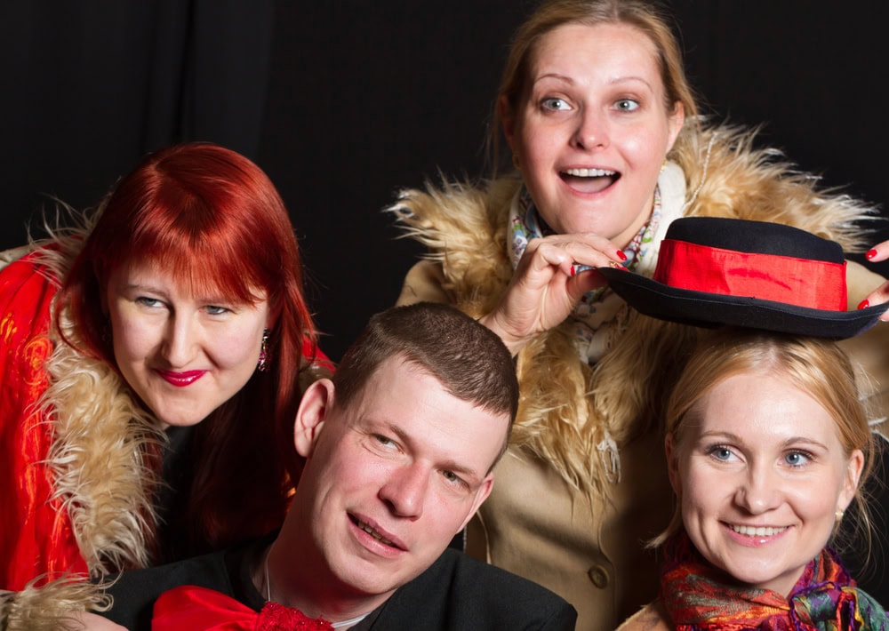 Four adults posing together, smiling. Two women wear red clothing, one holds a red and black hat above a man's head. All wear fur-trimmed coats against a dark background.