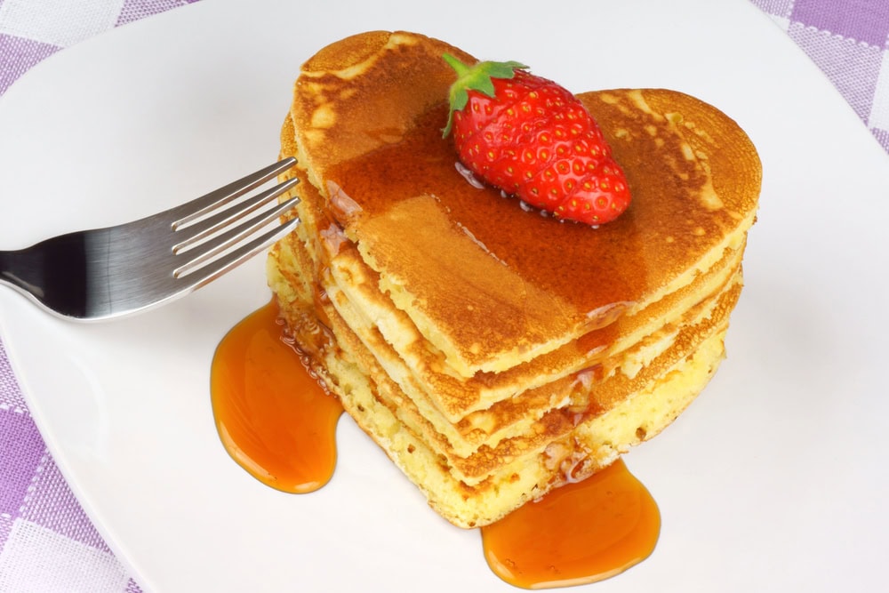 A stack of heart-shaped pancakes topped with a strawberry and syrup on a white plate, with a fork beside the stack.