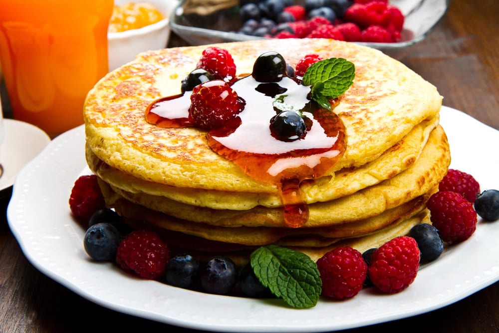 A stack of pancakes topped with syrup, berries, and mint leaves, served on a white plate with more berries around the side.