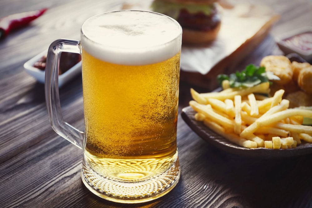 A glass mug of light beer with a frothy head sits on a wooden table next to a plate of French fries and another dish of food in the background.
