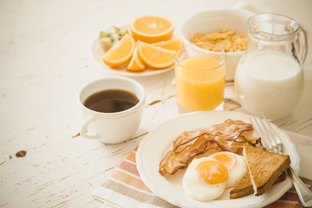A breakfast table with fried eggs, bacon, toast, a cup of coffee, a glass of orange juice, a jug of milk, cereal, and sliced oranges.