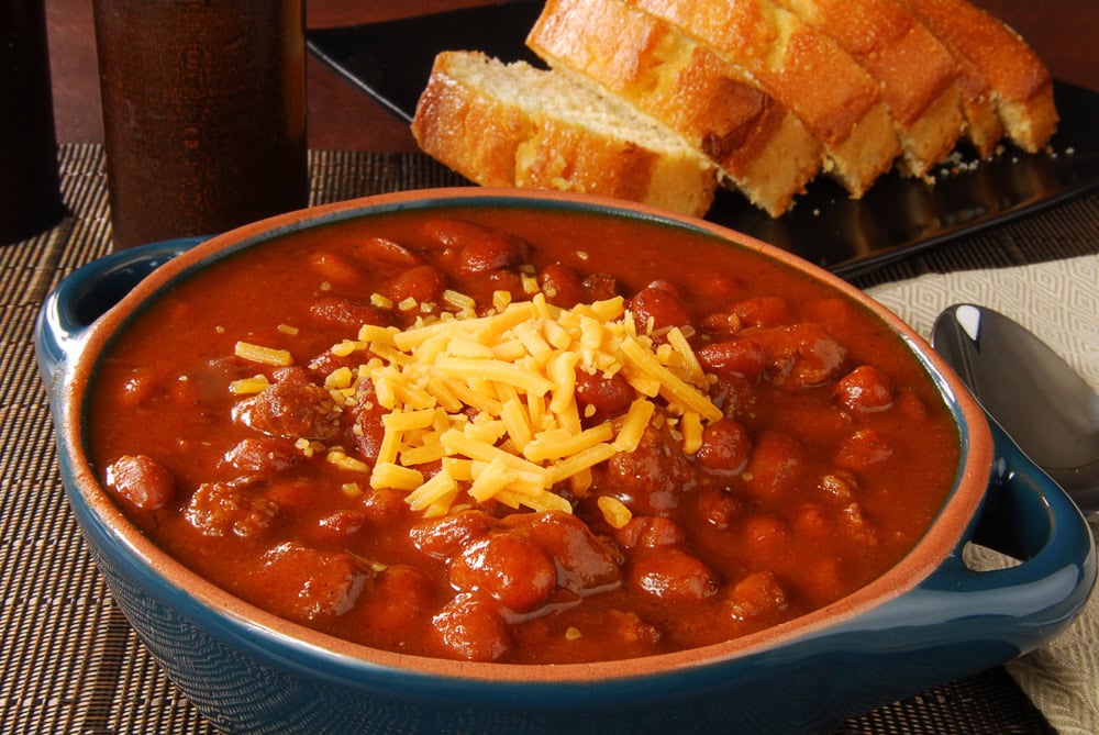 A bowl of chili topped with shredded cheddar cheese, served with sliced bread on a black plate in the background.