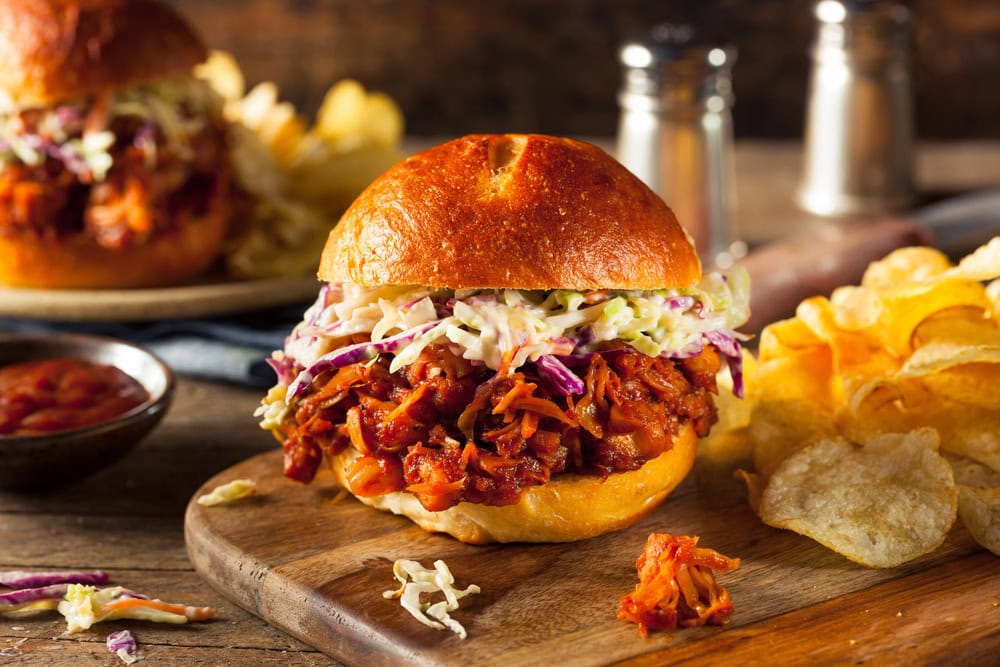 A pulled pork sandwich topped with coleslaw on a wooden board, served with potato chips; salt and pepper shakers and a plate are in the background.