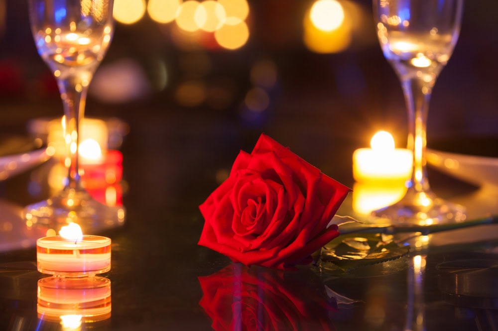 A red rose lying on a table surrounded by lit candles and two empty wine glasses, with a blurred background of warm lights.