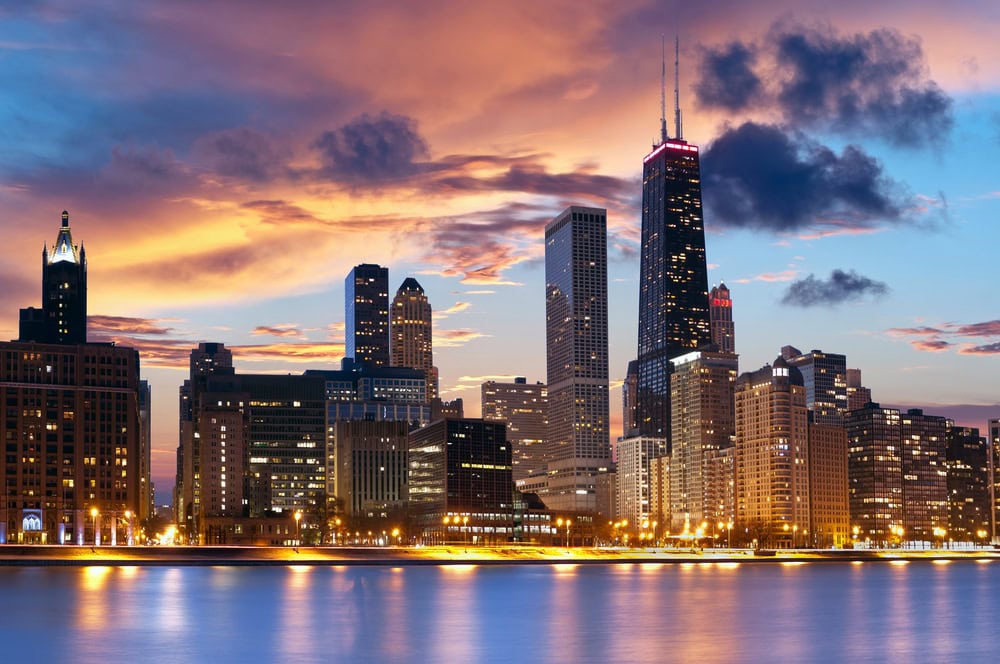 Chicago skyline at dusk with illuminated buildings, including the John Hancock Center, under a colorful sky, reflected in the calm water below.