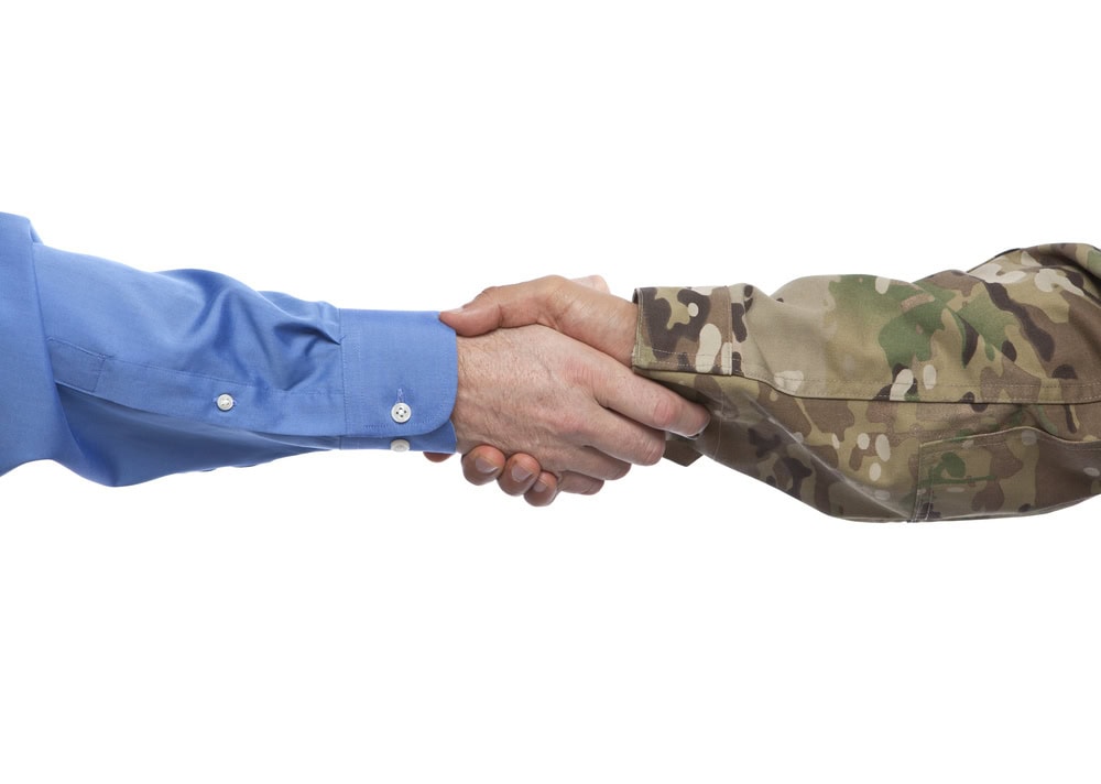A person in a blue dress shirt shakes hands with a person in a military camouflage uniform against a white background.