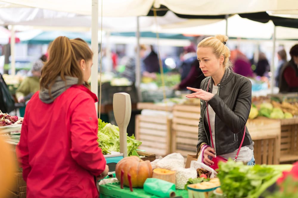 Two women interact at an outdoor market produce stand; one gestures while the other stands behind a table with vegetables and a scale.