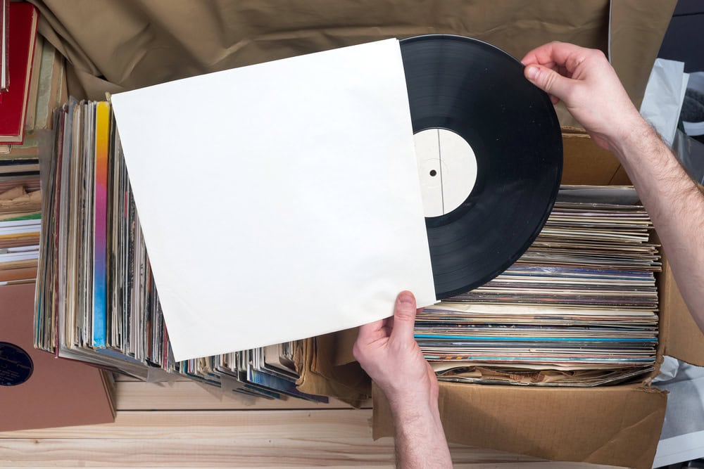 A person removes a vinyl record from a plain white sleeve above boxes filled with more vinyl records.