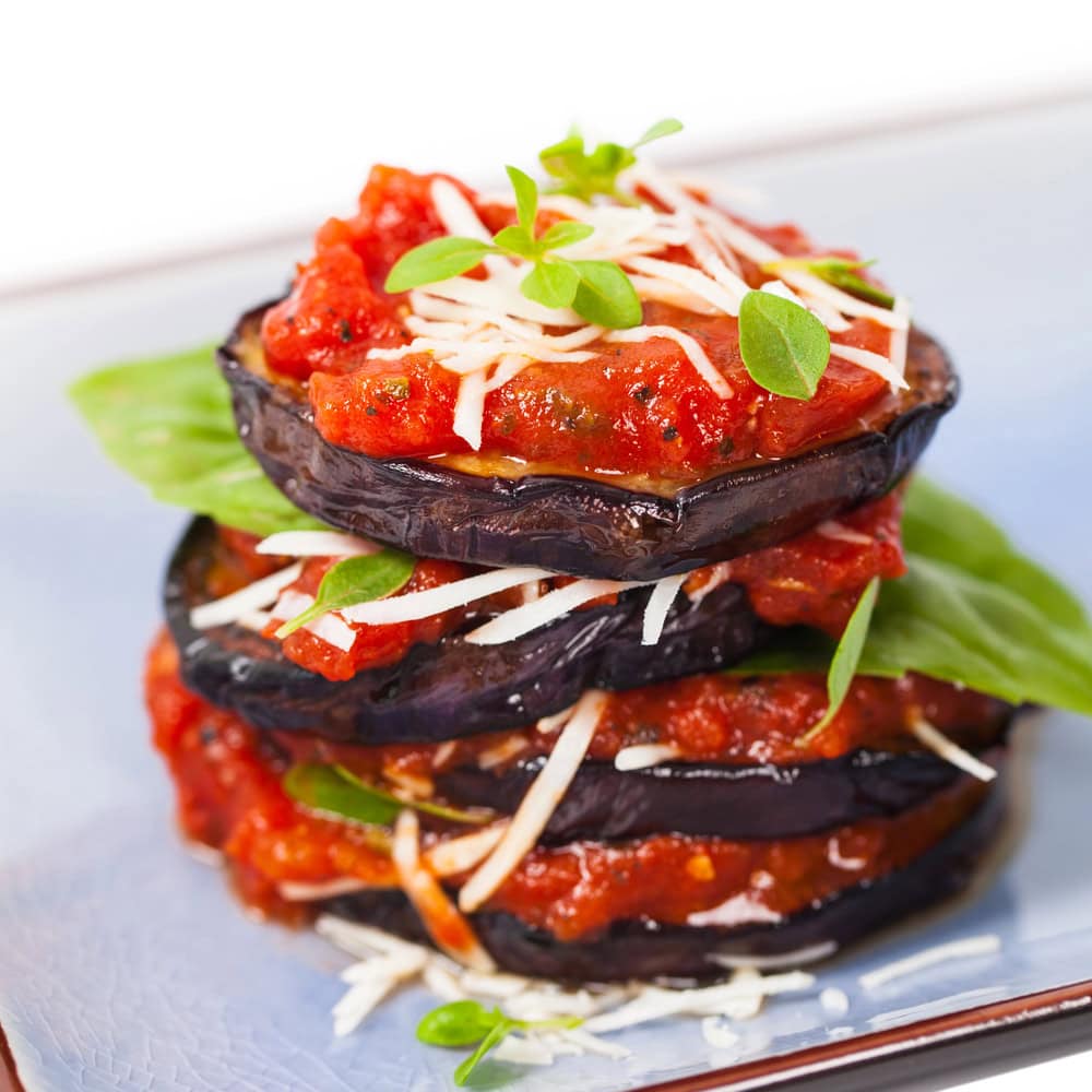 Stacked slices of roasted eggplant layered with tomato sauce, shredded cheese, fresh basil leaves, and garnished with small green herbs on a plate.