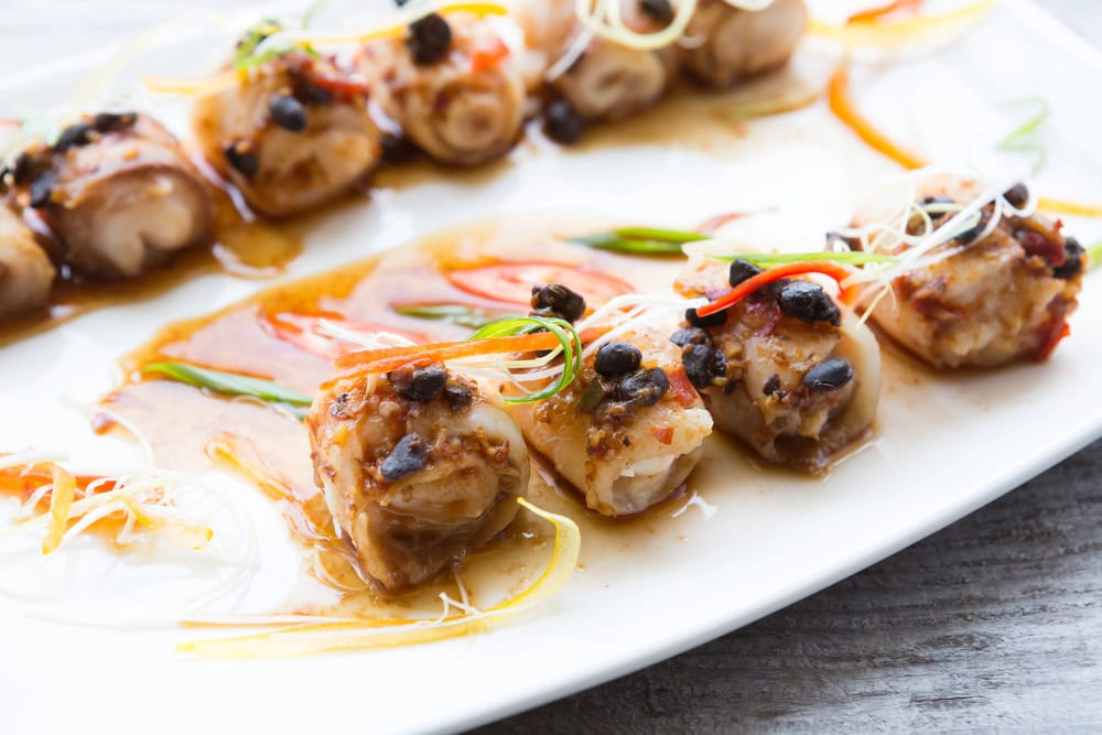 A white plate with several pieces of steamed fish rolls topped with black bean sauce, sliced chili, and garnished with thinly sliced vegetables.