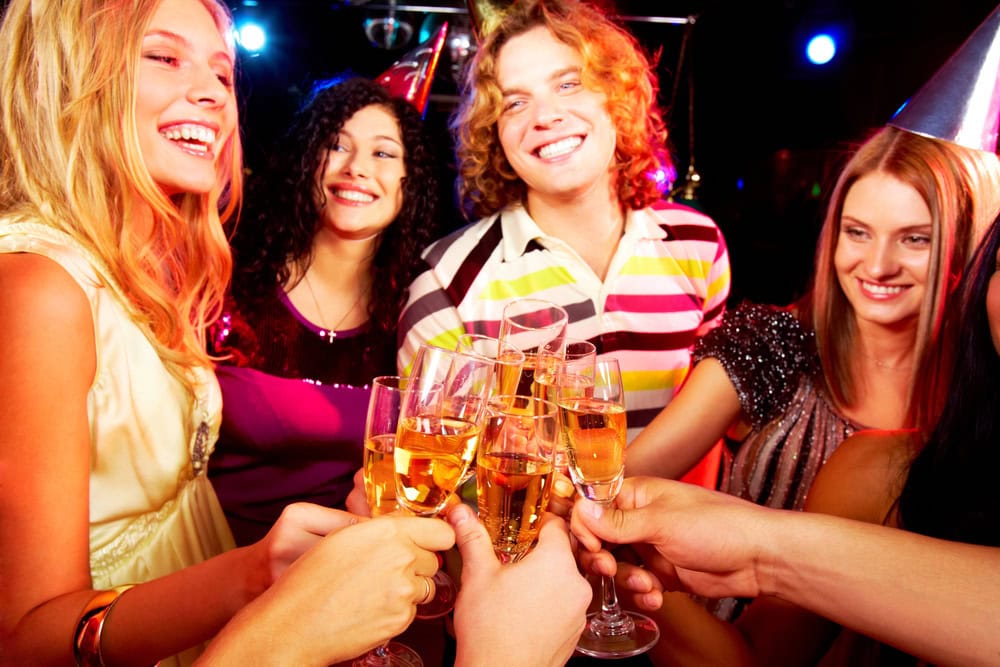A group of young adults in party attire clink glasses of champagne together, smiling and celebrating in a lively, colorful setting.