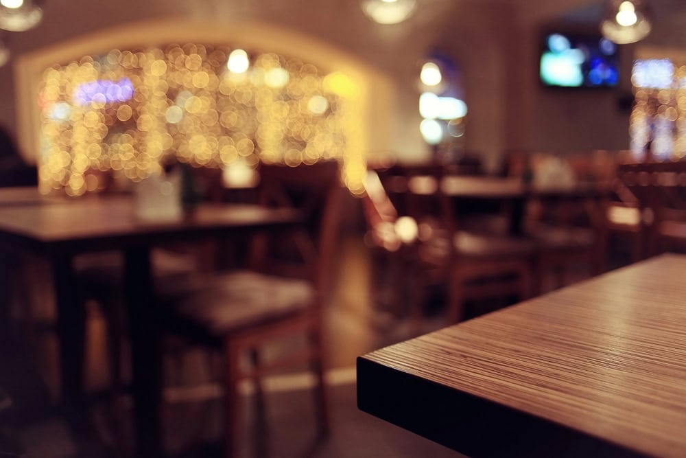 A close-up of a wooden table in an empty restaurant with blurred chairs, string lights, and a TV in the background.