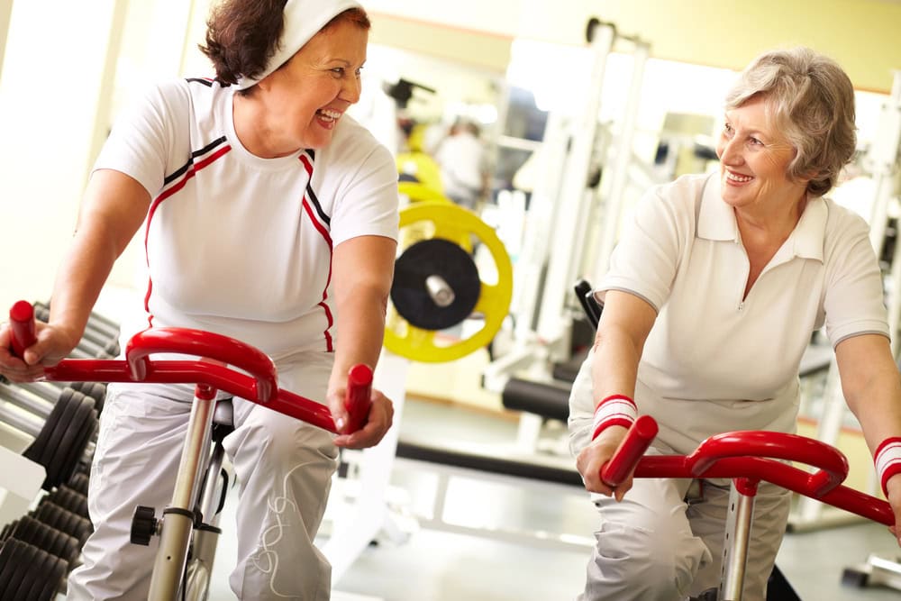 Two older women exercise on stationary bikes at a gym, smiling and talking to each other. Gym equipment and weights are visible in the background.