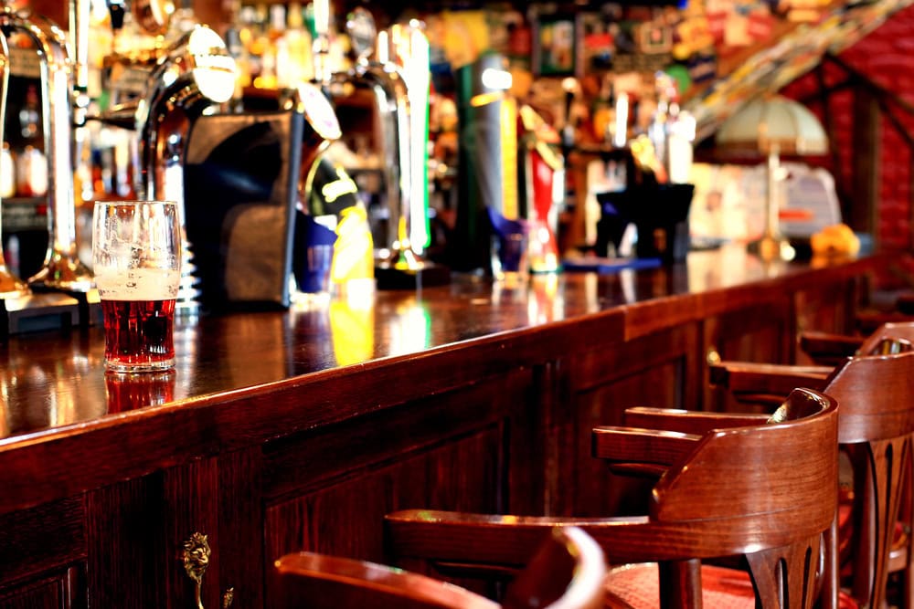 A wooden bar counter with a partially filled glass of beer, several beer taps, and empty wooden bar stools in a well-lit pub.