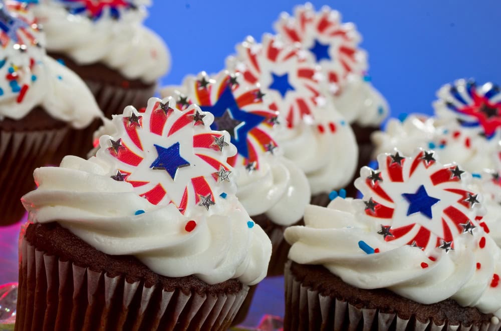 Chocolate cupcakes with white frosting, topped with red, white, and blue star decorations and sprinkles, arranged closely together.