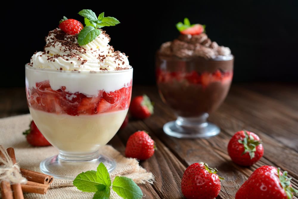 Two dessert parfaits in glass cups: one with vanilla pudding, strawberries, and whipped cream; the other with chocolate pudding, strawberries, and whipped cream, surrounded by fresh strawberries.