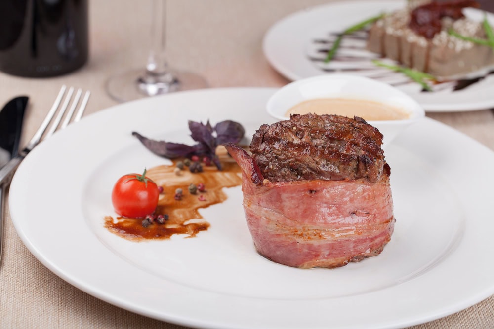 A bacon-wrapped filet mignon served on a white plate with a small tomato, sauce, herbs, and peppercorn garnish. Another plated dish is blurred in the background.