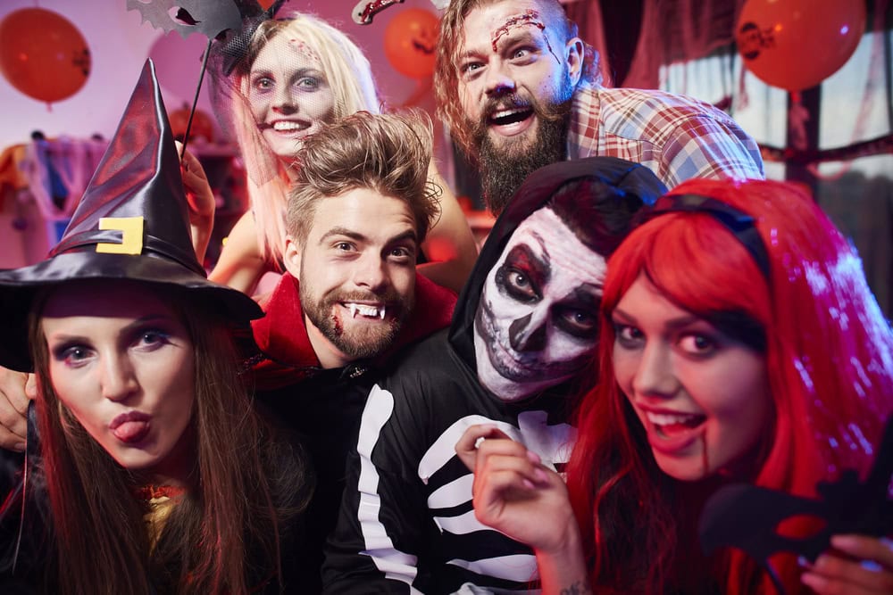 A group of six people in Halloween costumes, including a witch, skeleton, vampire, and red-haired character, pose together and smile at a festive indoor party.
