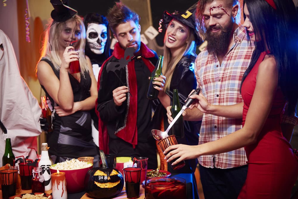 A group of people in Halloween costumes stand around a decorated table with drinks and snacks, socializing at a party.