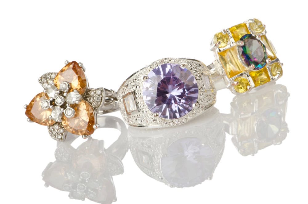 Three gemstone rings with intricate designs, featuring orange, purple, and multicolored stones set in silver metal, displayed on a reflective surface.