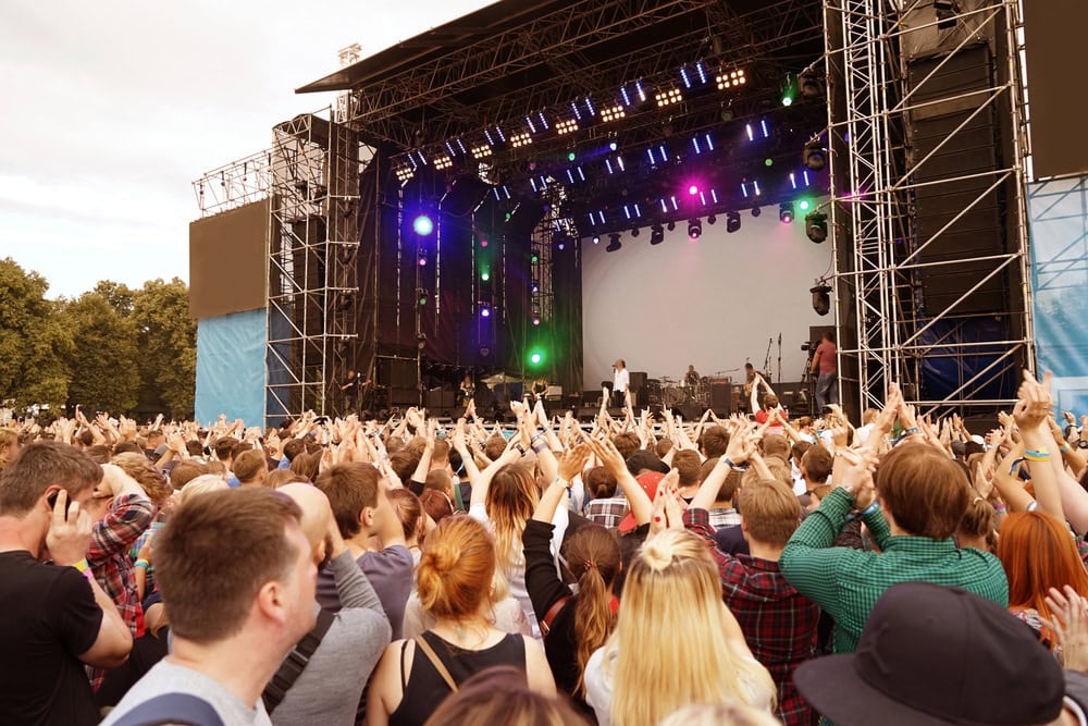Large crowd gathered in front of an outdoor concert stage with lights, musicians performing, and hands raised in the air.