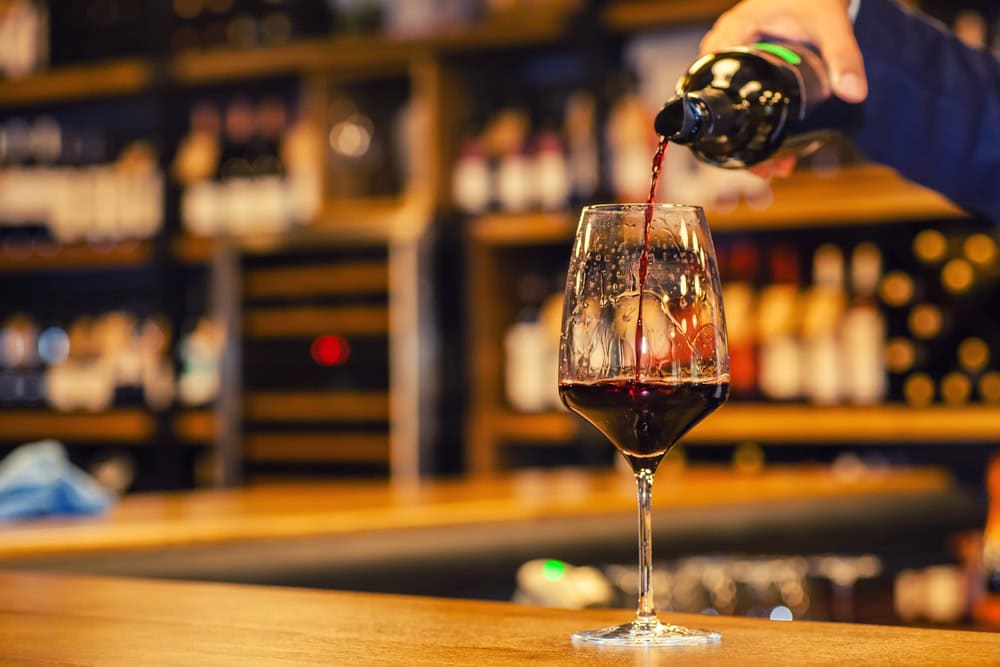 A hand pours red wine from a bottle into a glass on a wooden bar; shelves with bottles are blurred in the background.