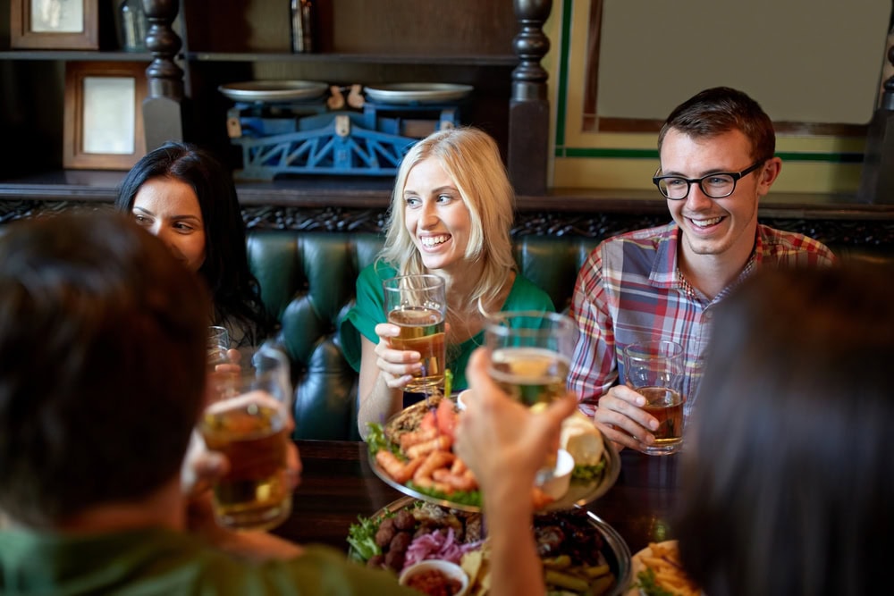 Five people sit around a table in a restaurant or pub, smiling and holding drinks, with plates of food in the center of the table.