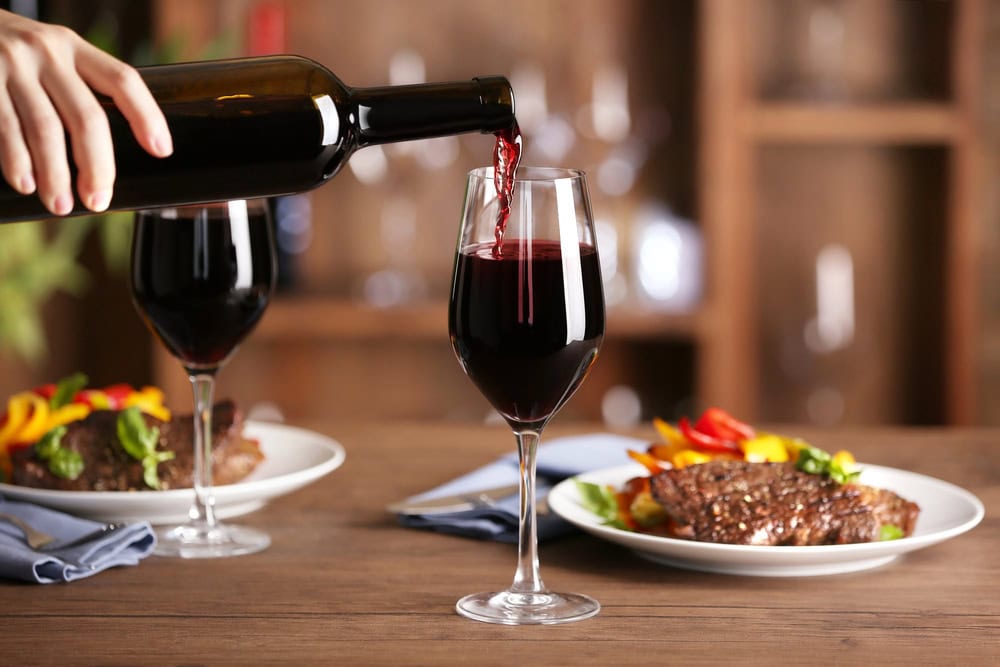 A hand pours red wine from a bottle into a glass on a wooden table set with plates of steak and vegetables.