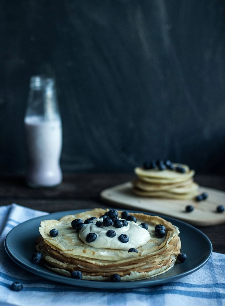 A stack of crepes topped with cream and blueberries on a dark plate, with more crepes and a bottle of milk in the background.