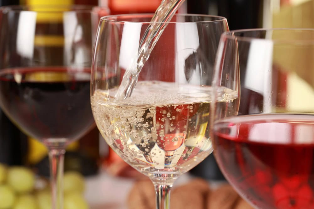 Close-up of three wine glasses: one being filled with white wine, one with red wine, and one with rosé, with blurred bottles and grapes in the background.