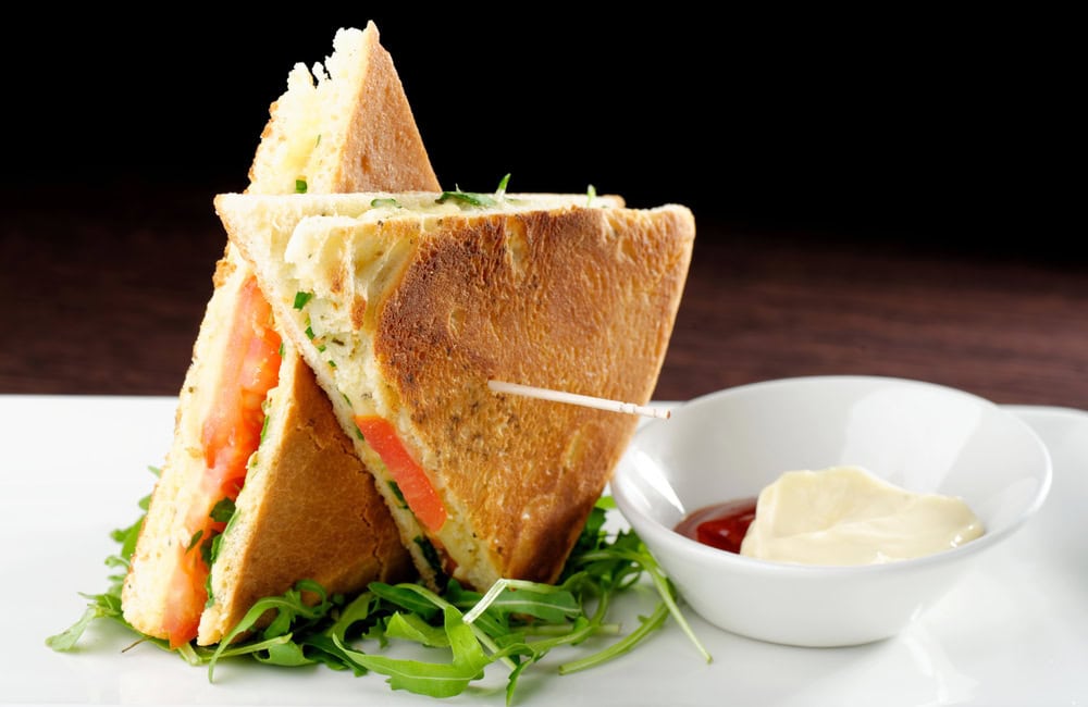 Two triangular sandwich halves with tomato, greens, and cheese on a white plate, served with a side of mayonnaise and ketchup in a small bowl.
