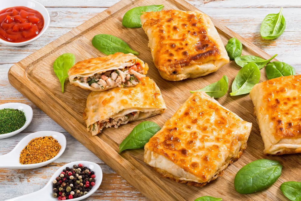 Five golden-brown stuffed pastries on a wooden board with fresh spinach leaves, surrounded by small bowls of spices and a dish of ketchup.