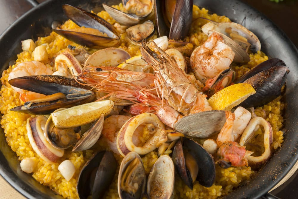 A close-up of a seafood paella, featuring yellow rice, shrimp, mussels, clams, squid rings, and lemon wedges in a black pan.