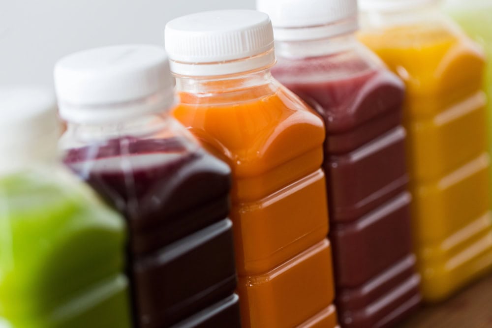 Five plastic bottles filled with different colored juices are lined up closely together, each with a white cap.