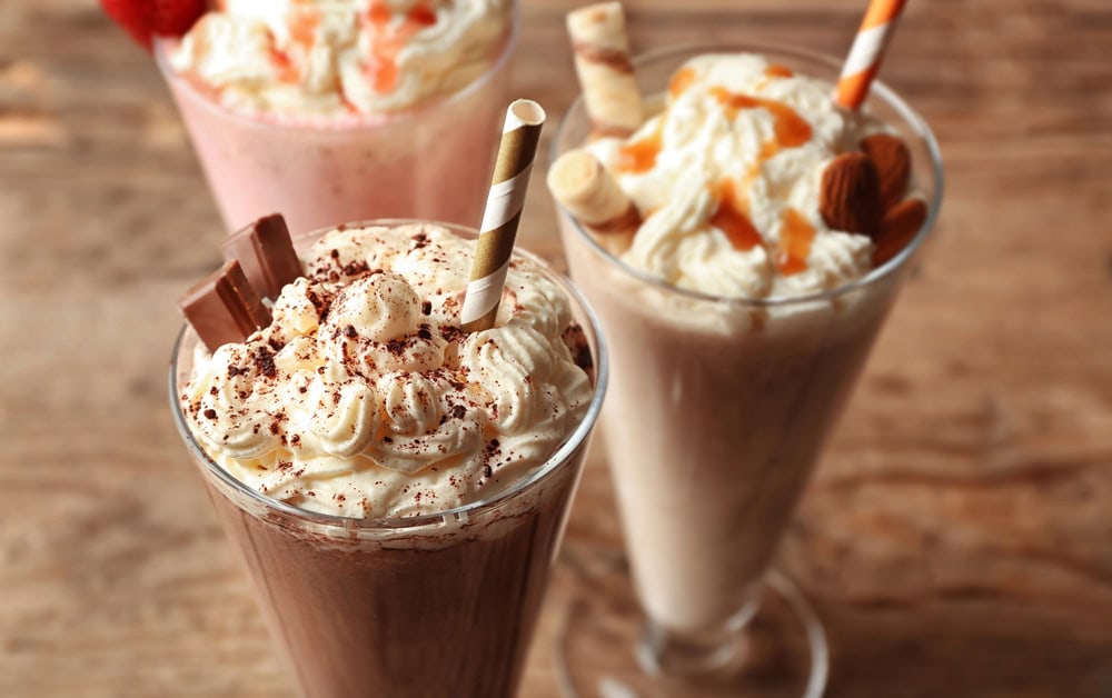 Three milkshakes in tall glasses topped with whipped cream, chocolate pieces, caramel drizzle, almonds, and wafer rolls, placed on a wooden surface.