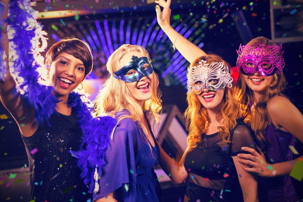 Four women wearing colorful masquerade masks and dresses pose together, smiling and celebrating amid confetti at a festive party.