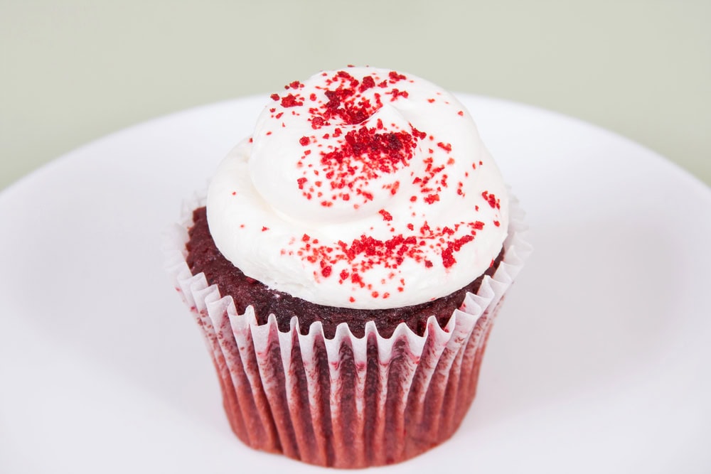 A red velvet cupcake with white frosting and red sprinkles is placed on a white plate.
