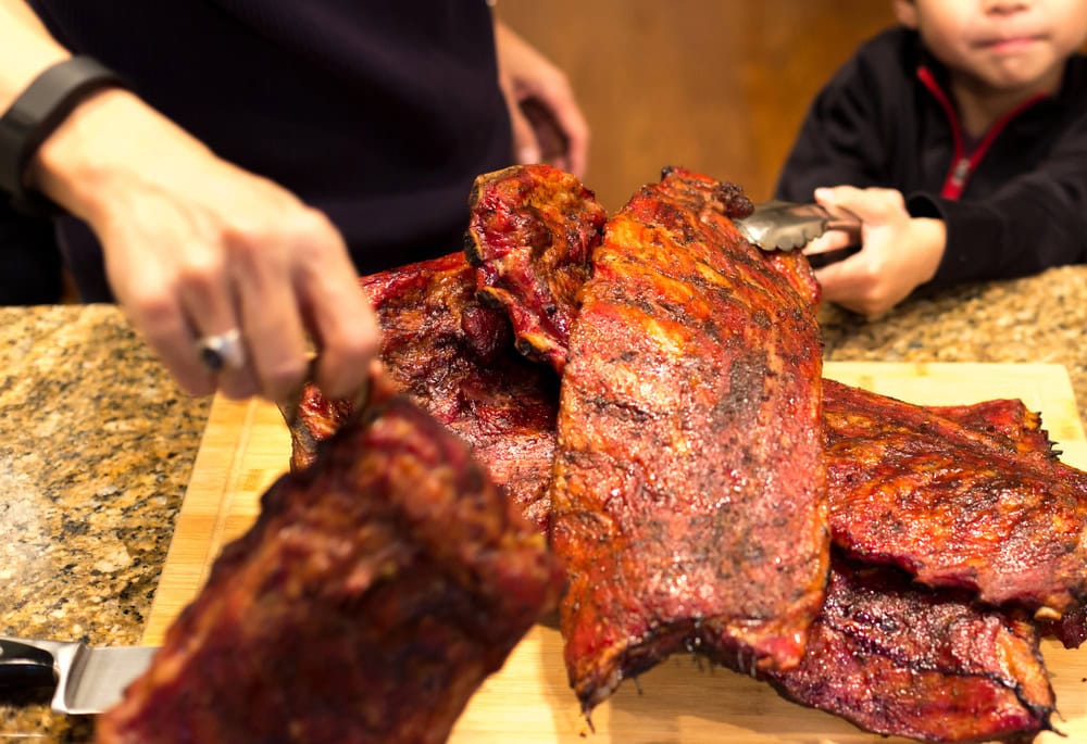 Two people, one adult and one child, hold utensils as they serve several racks of barbecued ribs on a wooden cutting board.