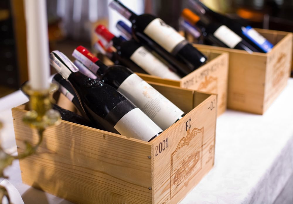 Several wine bottles with blank labels are arranged in three wooden crates on a table with a white tablecloth.