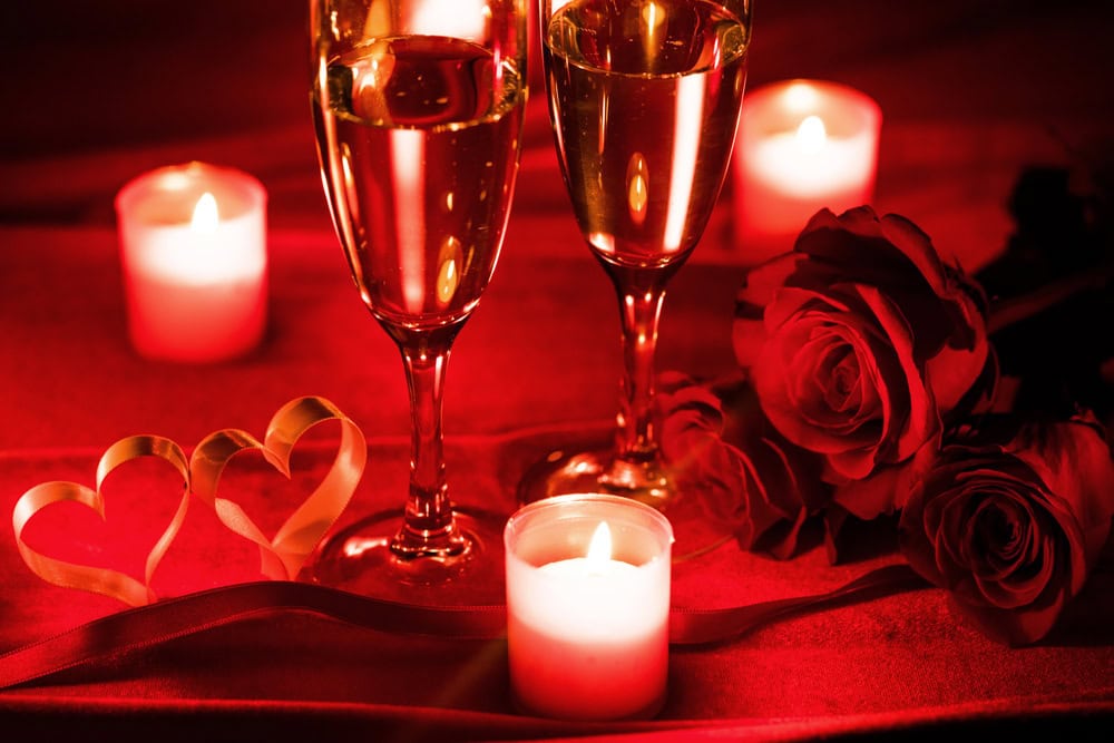 Two glasses of champagne, red roses, lit candles, and two ribbon hearts are arranged on a red surface, creating a romantic atmosphere.