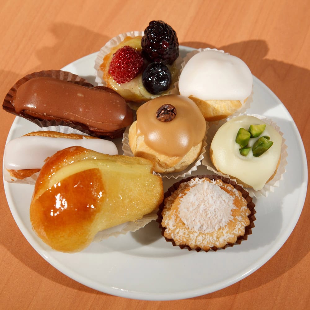 A white plate with assorted Italian pastries, including cream puffs, fruit tarts, and éclairs, on a wooden table.