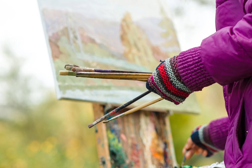 A person in a purple jacket and striped gloves holds paintbrushes while painting on a canvas outdoors.