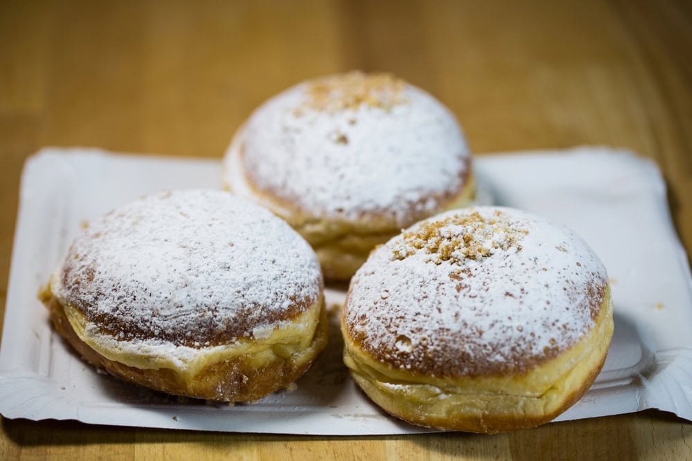 Three round, powdered sugar-covered jelly donuts sit on a white tray atop a wooden surface.