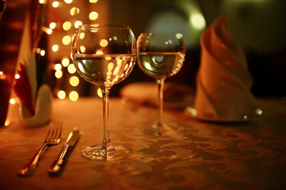 Two wine glasses filled with water sit on a decorated table with a fork, knife, napkin, and warm string lights in the background.